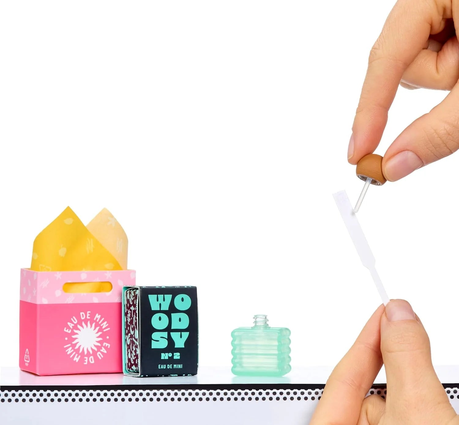 Hands placing a mini fragrance tool beside a pink box and teal mini bottle