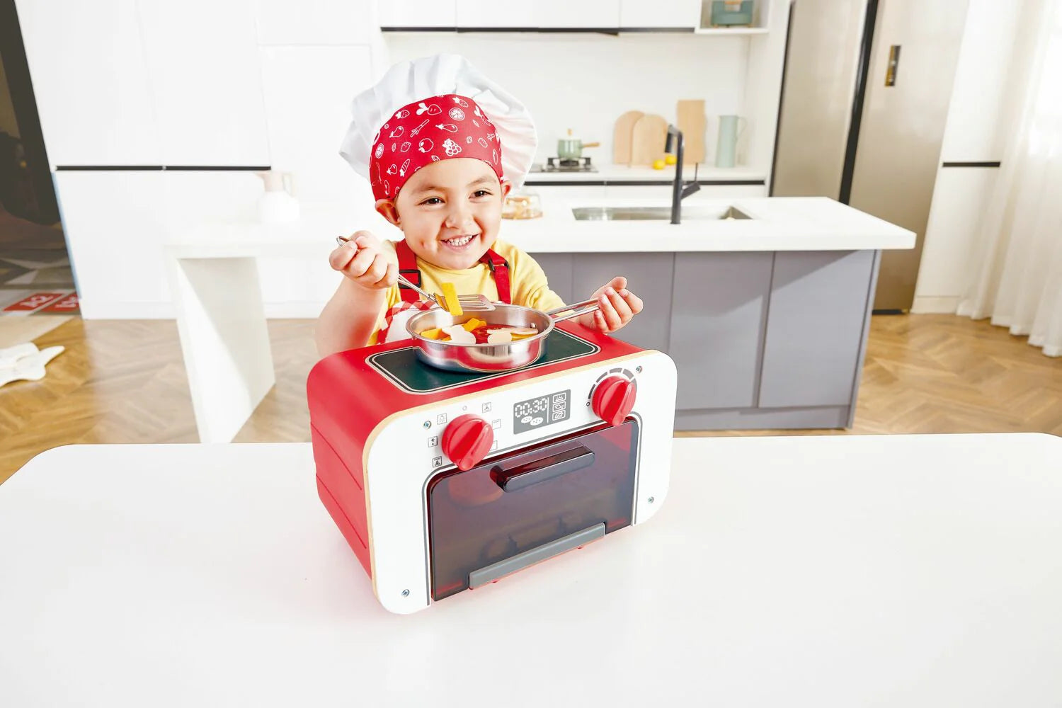 Child wearing a red chef hat playing with a red toy baking oven and cookie tray