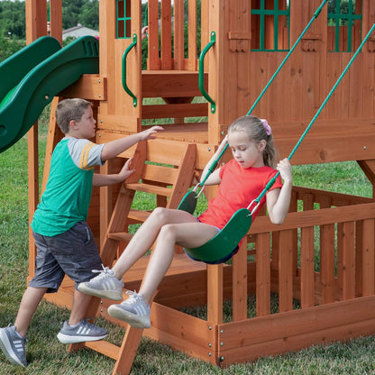 Kinder sitzen auf der Schaukel zwischen Holzpfosten am Spielset mit grüner Rutsche