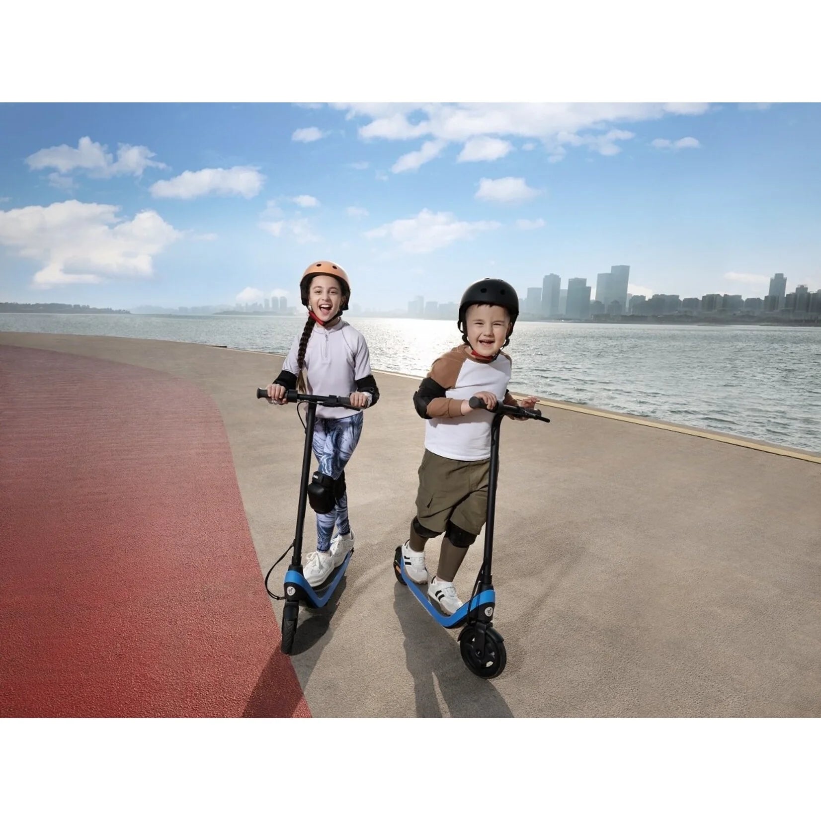 Two kids riding blue kick scooters with black wheels on a waterfront walkway