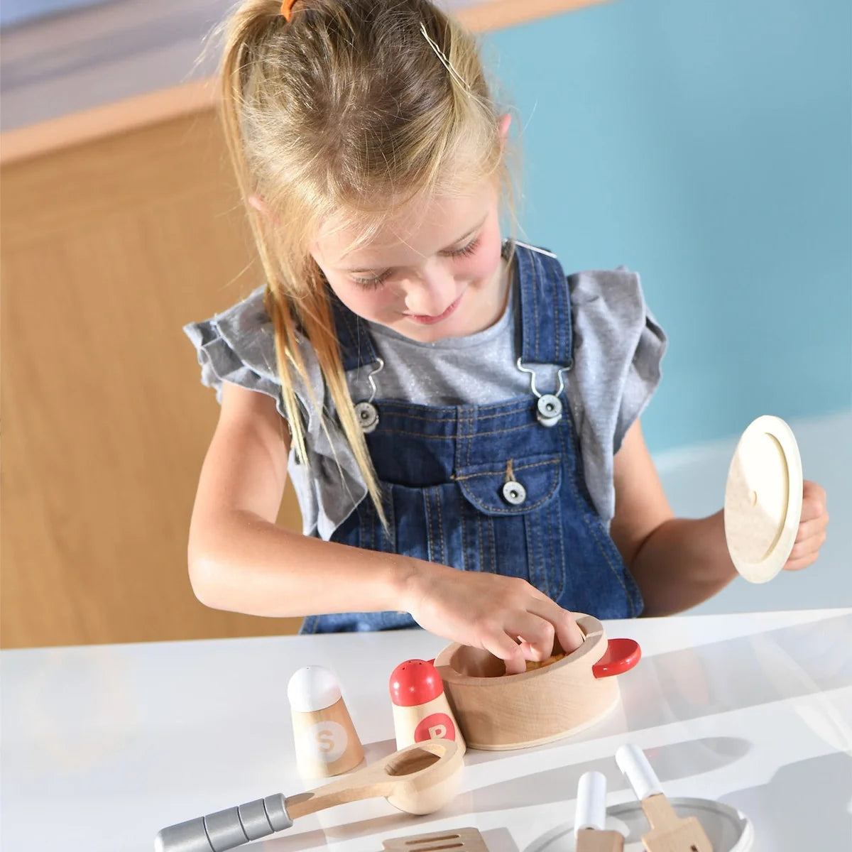 Kids cook set in use: wooden pot with red lid knob, spoon, and plates near child