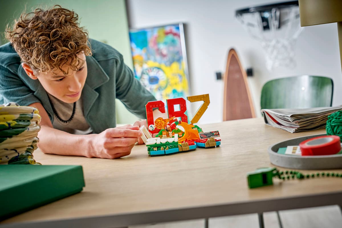 Person assembling LEGO soccer highlights set at a wooden desk with scattered bricks