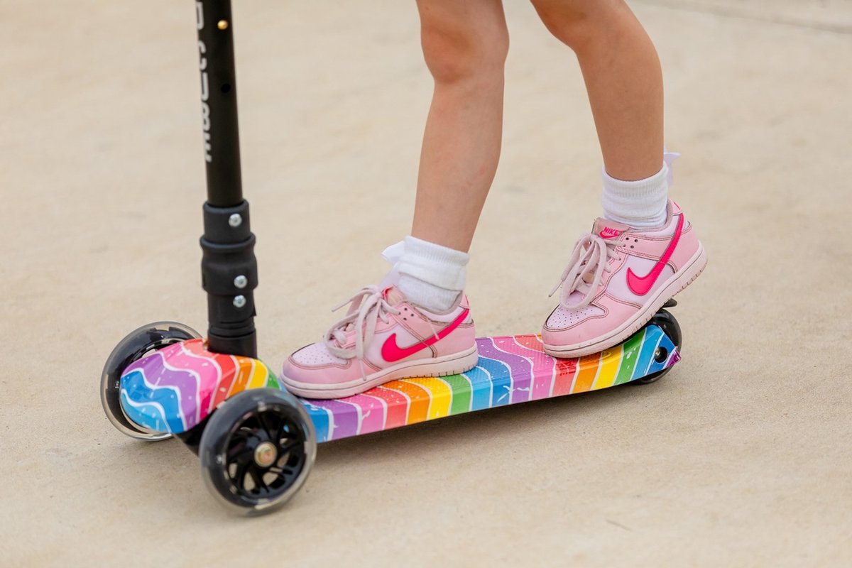 Kids' 3-wheel scooter with rainbow deck under pink sneakers; wheels visible.