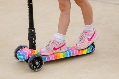 Kids' 3-wheel scooter with rainbow deck under pink sneakers; wheels visible.