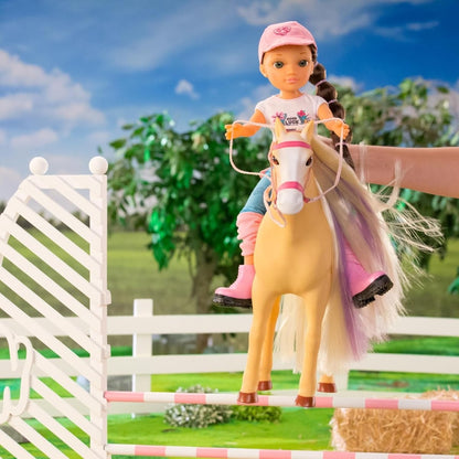 Doll with pink cap on light tan horse beside fence; white flower shirt and jeans